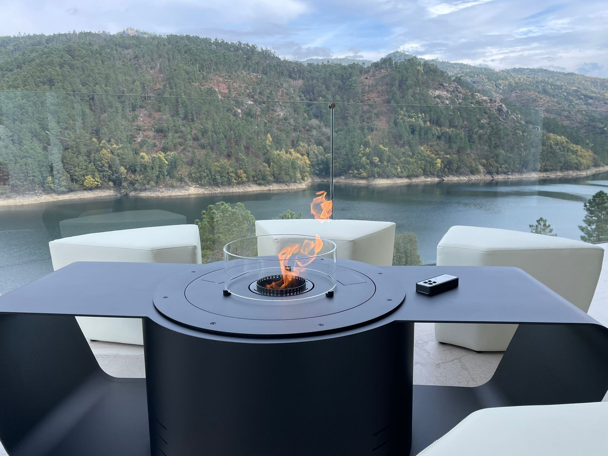 Circus with automated bioethanol burner and white ottomans. Close up on the remote control and panoramic view on the river, the hills and the woods in the background