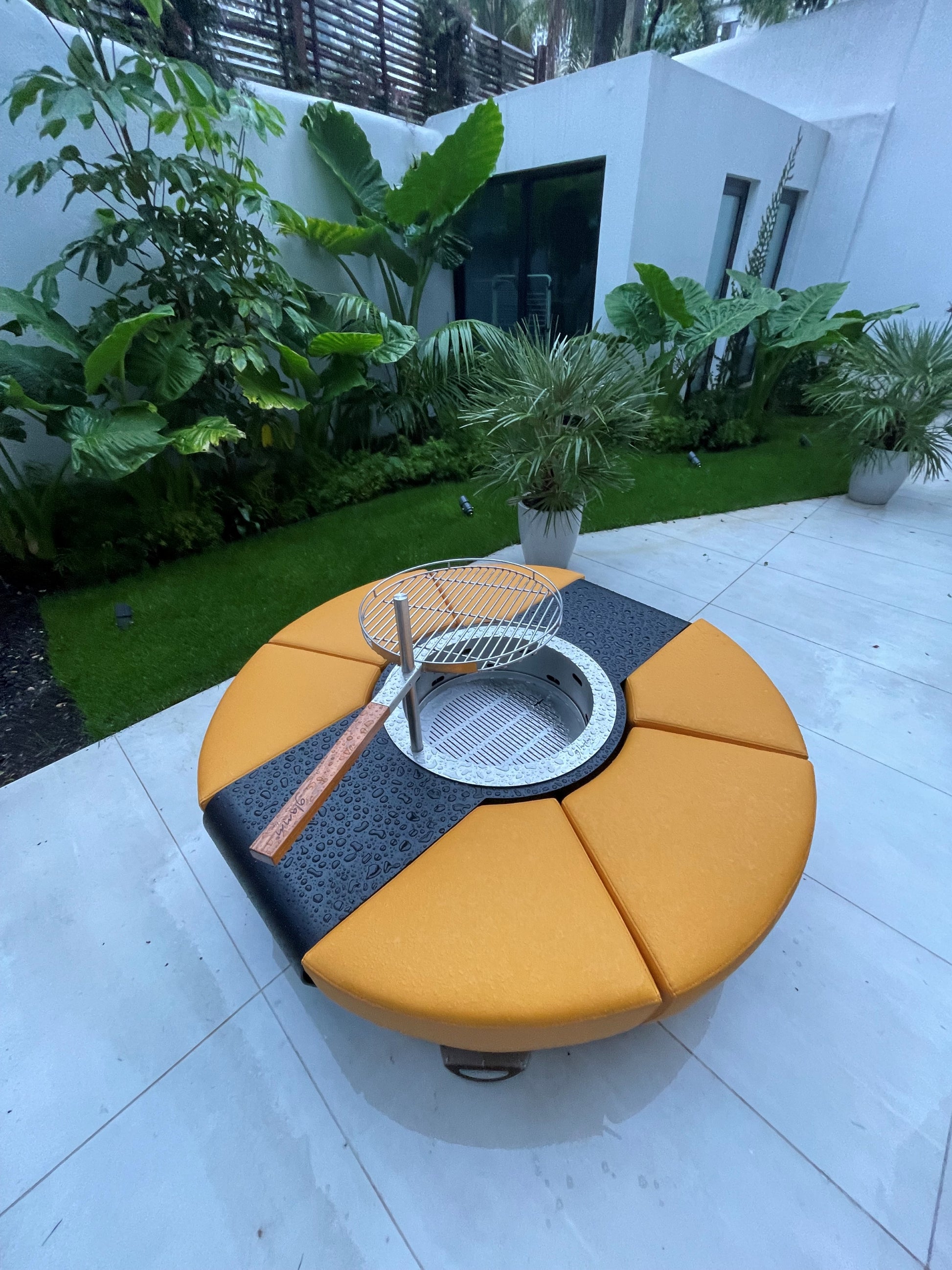 Circus with wood burner, barbecue grill and saffron yellow ottomans in palm garden. The firepit is off and wet from rain.